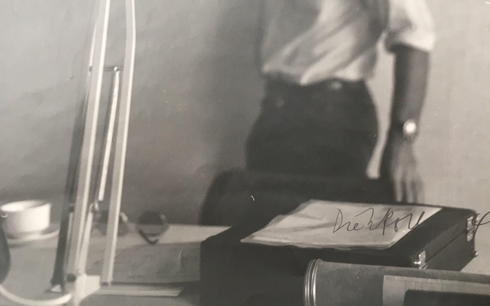 Eine Schwarz Weiß Fotographie zeigt im Vordergrund scharf eine Schreibtischlampe. Im Hintergrund sind man unscharf einen Mann mit Bart und einem weißen Hemd. Es ist Hansjörg Mayer.