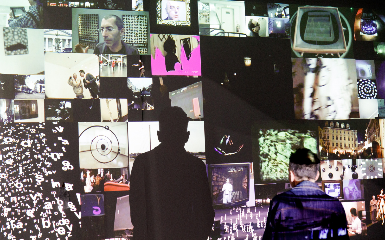 Zu sehen ist eine Video-Installation. Es werden viele kleinere Bilder auf eine Wand projiziert, wobei ein Mosaikartiges Bild entsteht. Davor steht ein betrachtender Mann. Er wirft einen auffälligen Schatten auf die Wand.