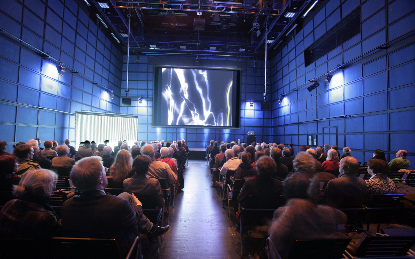 The ZKM_Media Theater full of people who look at a canvas which shows amorphous structures in white