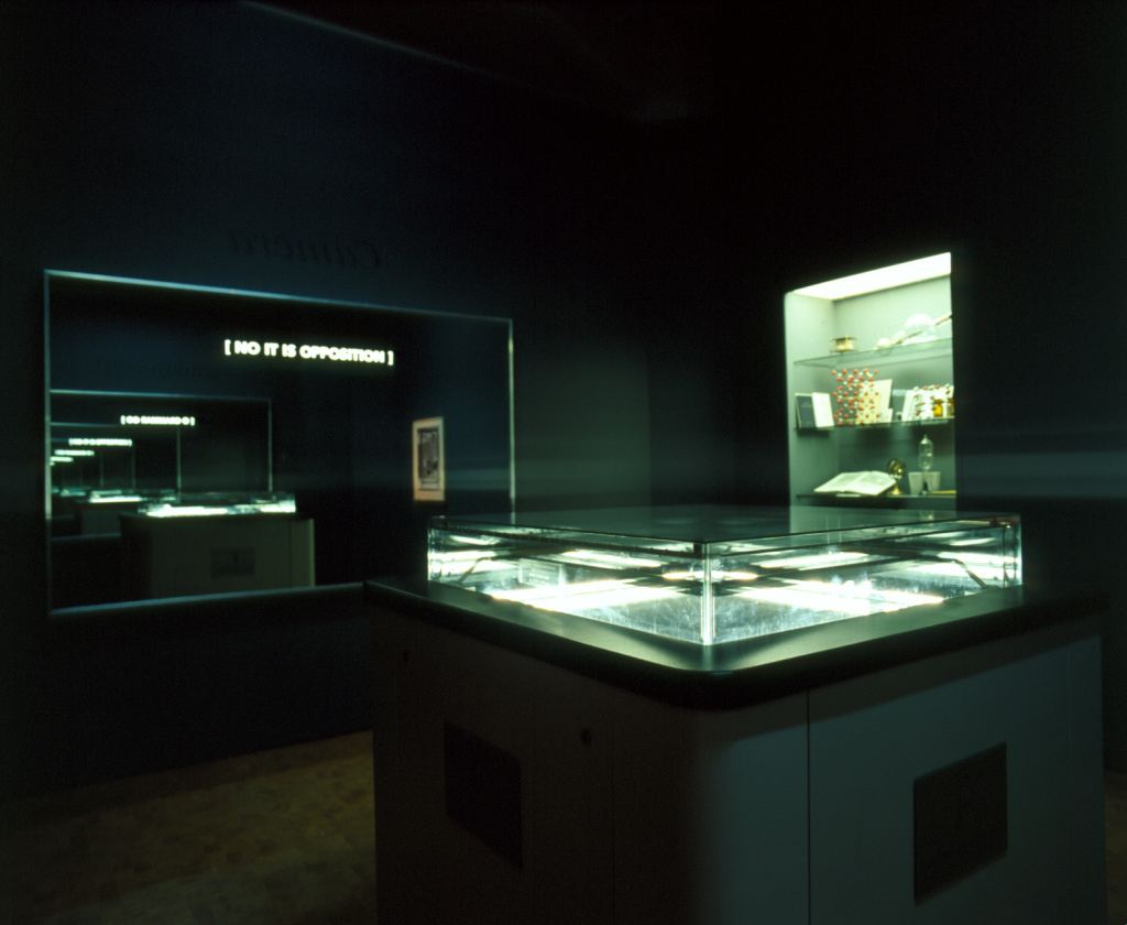 Der Blick in die Ausstellung »Iconoclash« zeigt "the typosophic pavilion"  von Ecke Bonk  und Richard Hamilton: einen dunklen Raum mit erleuchteten Vitrinen und einem Spiegel mit der Aufschrift »No It Is Opposition«
