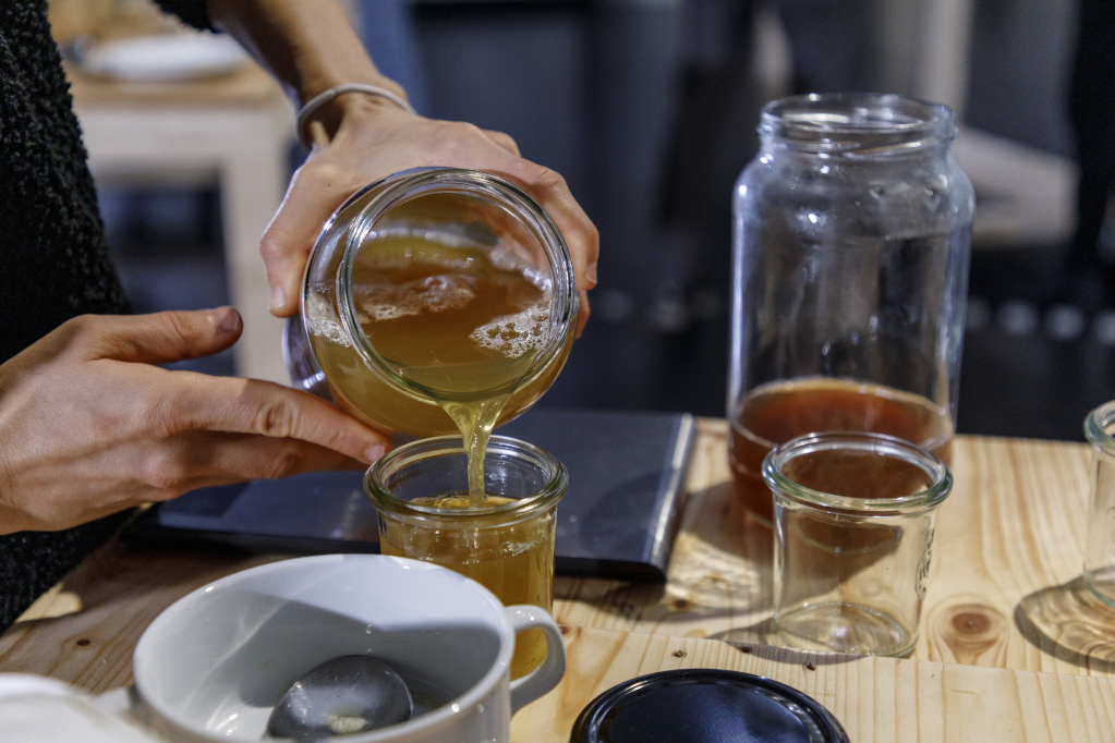 Aus einem Einmachglas wird eine Flüssigkeit in ein Glas auf einem Tische gefüllt. Auf dem Tisch stehen weitere Gläser und Schüsseln.