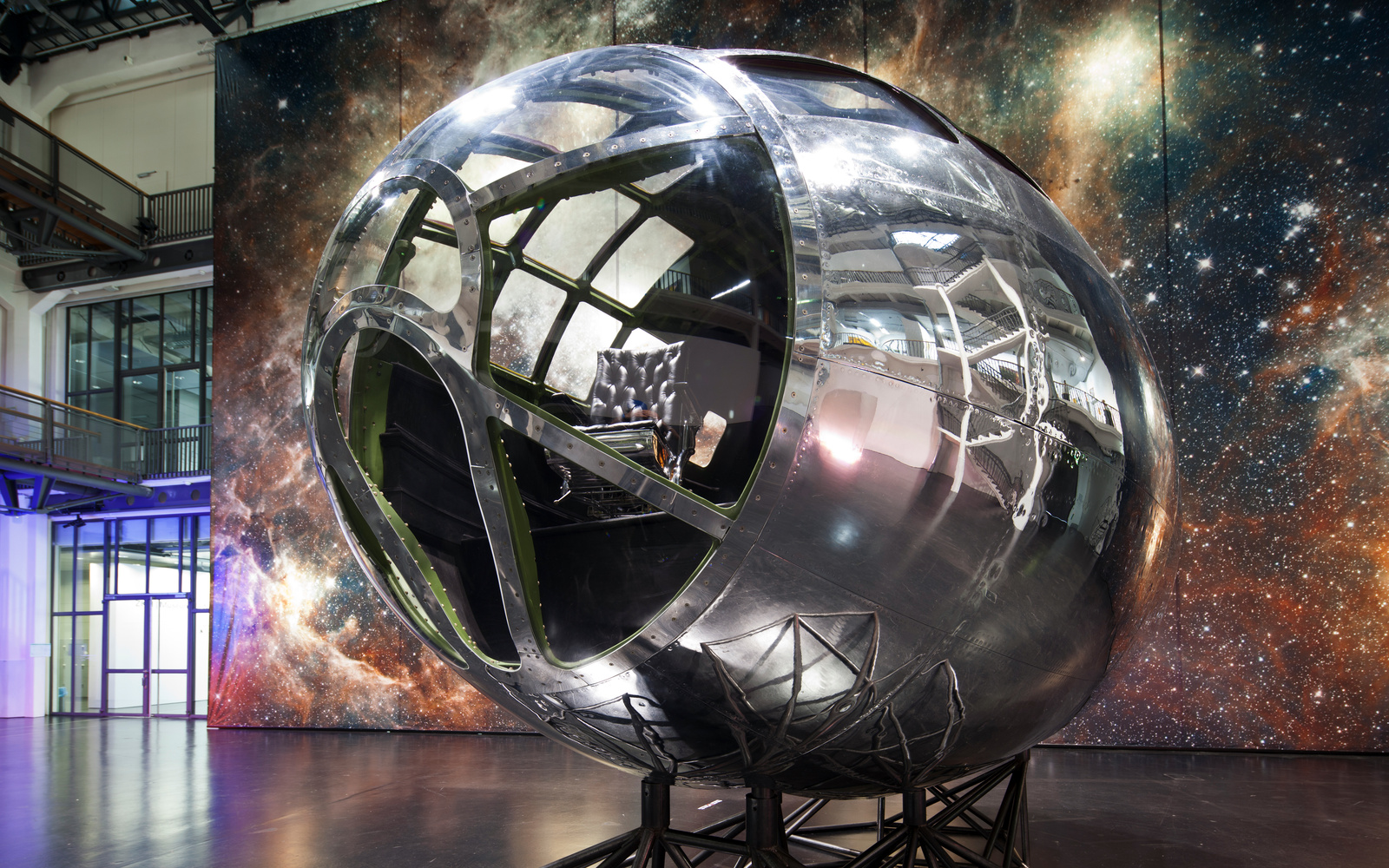 Zu sehen ist ein silbernes Gestellt, das an eine Raketenspitze erinnert und vor einer großen Leinwand mit dem Motiv einer Galaxie steht. In der vorderen Glaskuppel des Gestellt ist ein schwarzer Ledersessel zu erkennen. 