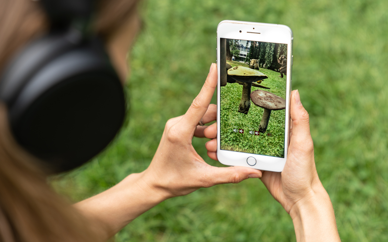 Zu sehen ist der Ausschnitt einer jungen Frau auf einer Wiese, die ein Handy in der Hand hat, auf dessen Screen Pilze auf der Wiese erscheinen.