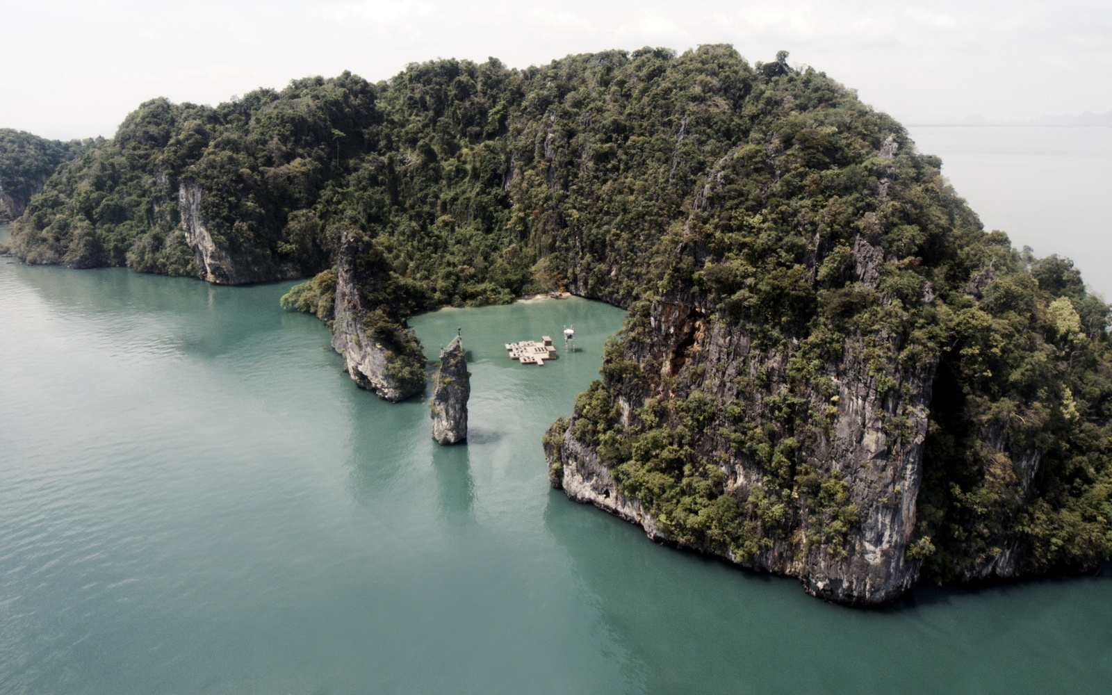 Das Bild zeigt eine von Wasser umgebene kleine Insel. In der Mitte des Bildes ist eine kleine Bucht. In dieser Bucht liegt ein Plateau mit zwei kleinen Bauten darauf, das auch rundherum von Wasser umgeben ist.