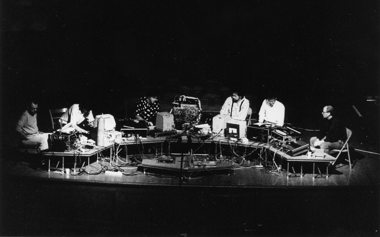 A black and white photo of the musician group »The Hub« from 1989, they are sitting in front of a long table with tube screens and many cables.