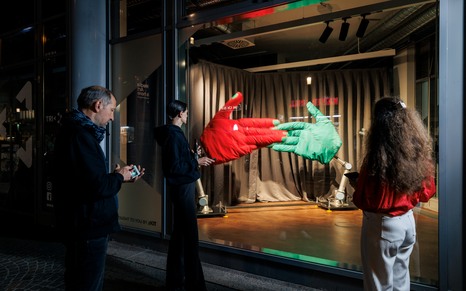 Auf dem Bild ist die Instalation »Handshake«,  AATB (Andrea Anner, Thibault Brevet), im Triangel am Kronenplatz zu sehen. Eine große ror und große grüne Hand sind am Boden fixiert und schlagen sich gegenseitig ein. Mehrere Menschen stehen vor der Scheibe.