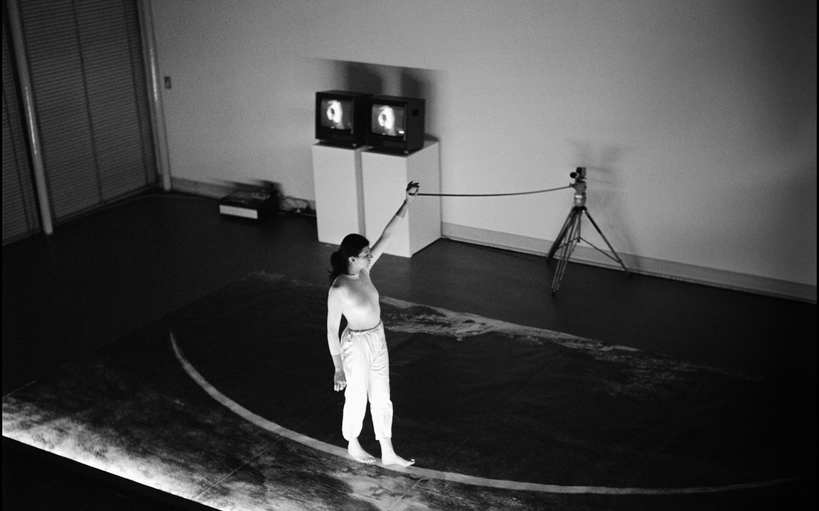 Ulrike Rosenbach, »Die einsame Spaziergängerin«, 1979. Zu sehen ist eine Person auf einer Bühne, die barfüßig auf einer gebogenen Linie läuft. Dabei hält sie ein Seil in der Hand, das an einem Stativ befestigt ist. Das Bild ist schwarz-weiß. 