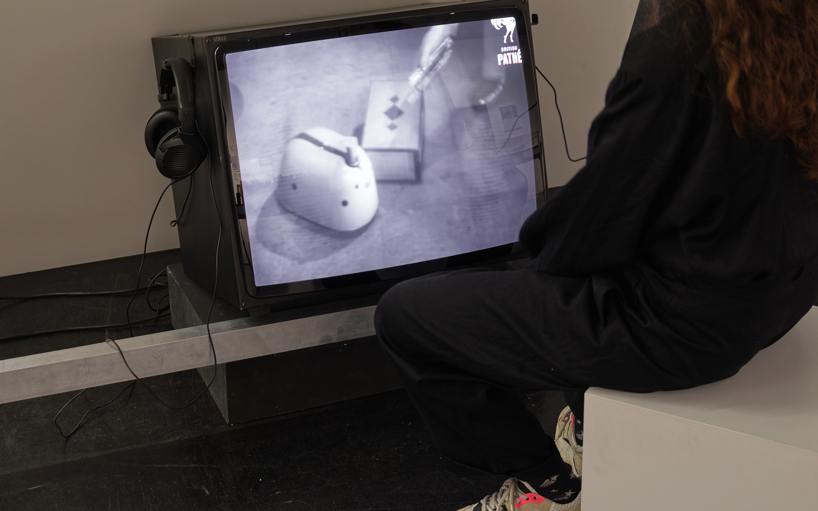 »ELMER – (ELectro-MEchanical Robot)« von William Grey Walter. Zu sehen ist eine Person, die auf einen schwarz-weiß Röhrenfernseher schaut. Sie trägt Kopfhörer und sitzt auf einem weißen Hocker.
