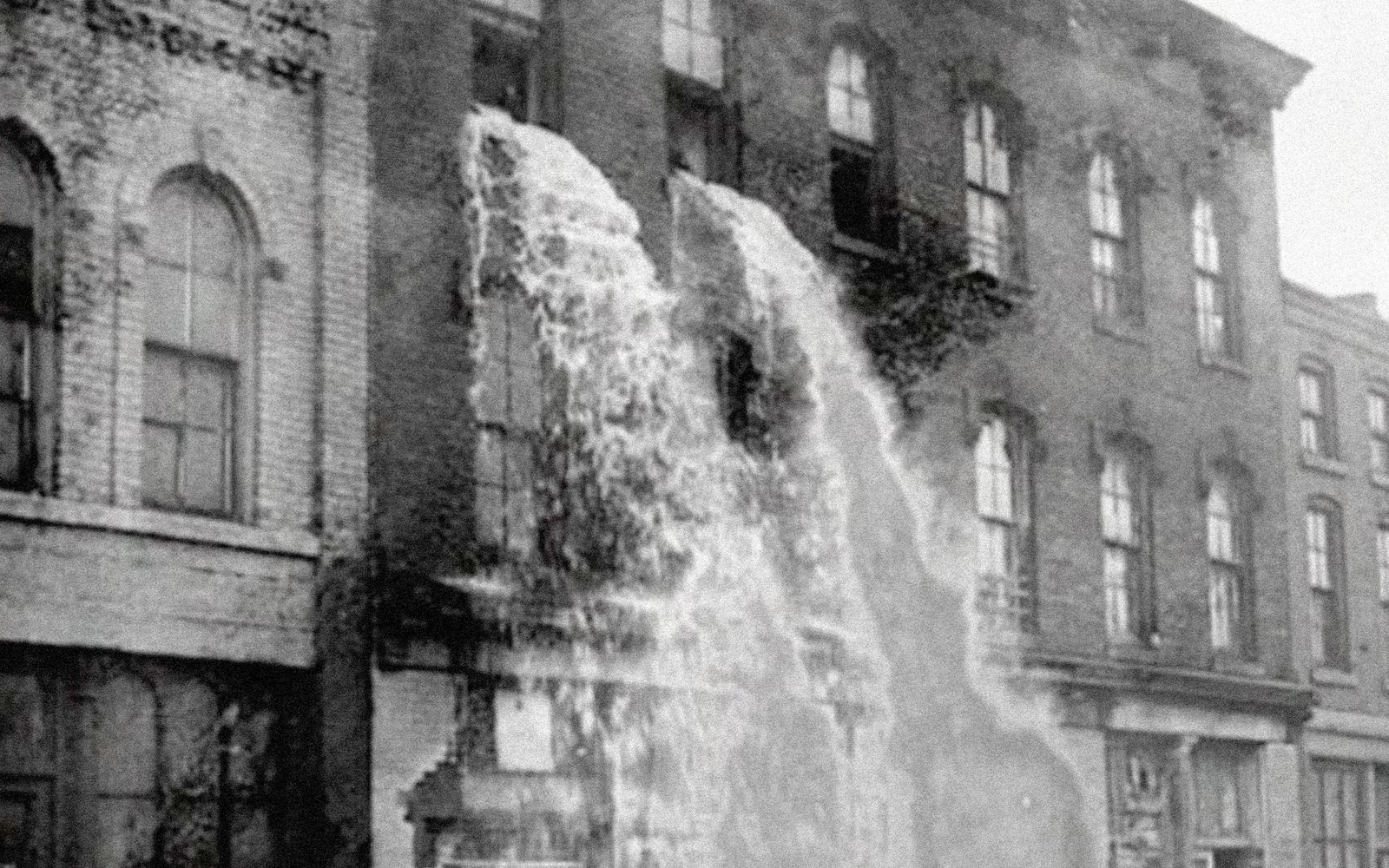 Man sieht ein Fabrikgebäude, aus dessen Fenster ein Wasserfall hervorbricht in Schwarz-Weiß.