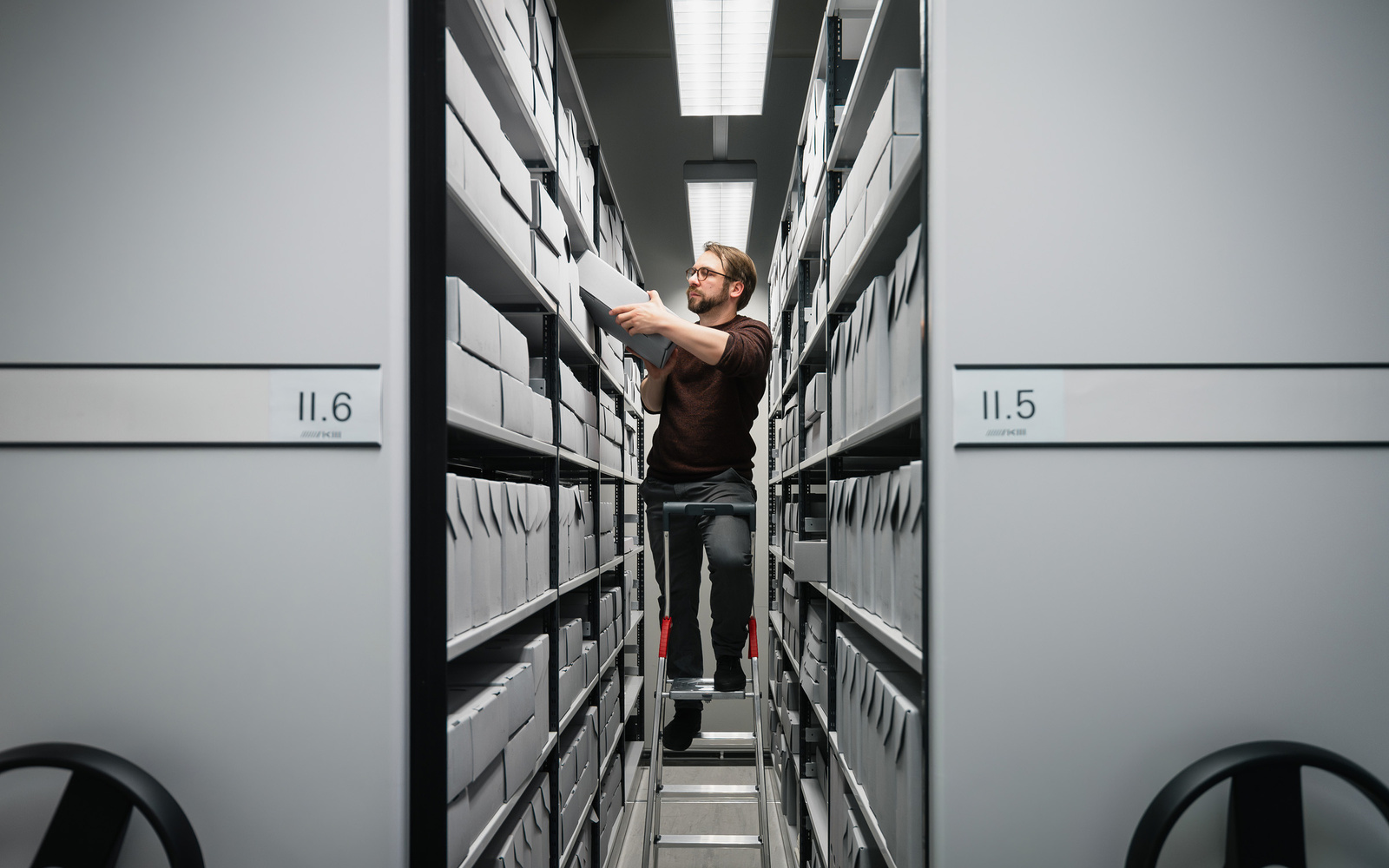 One person stands on a ladder and puts away archive boxes.
