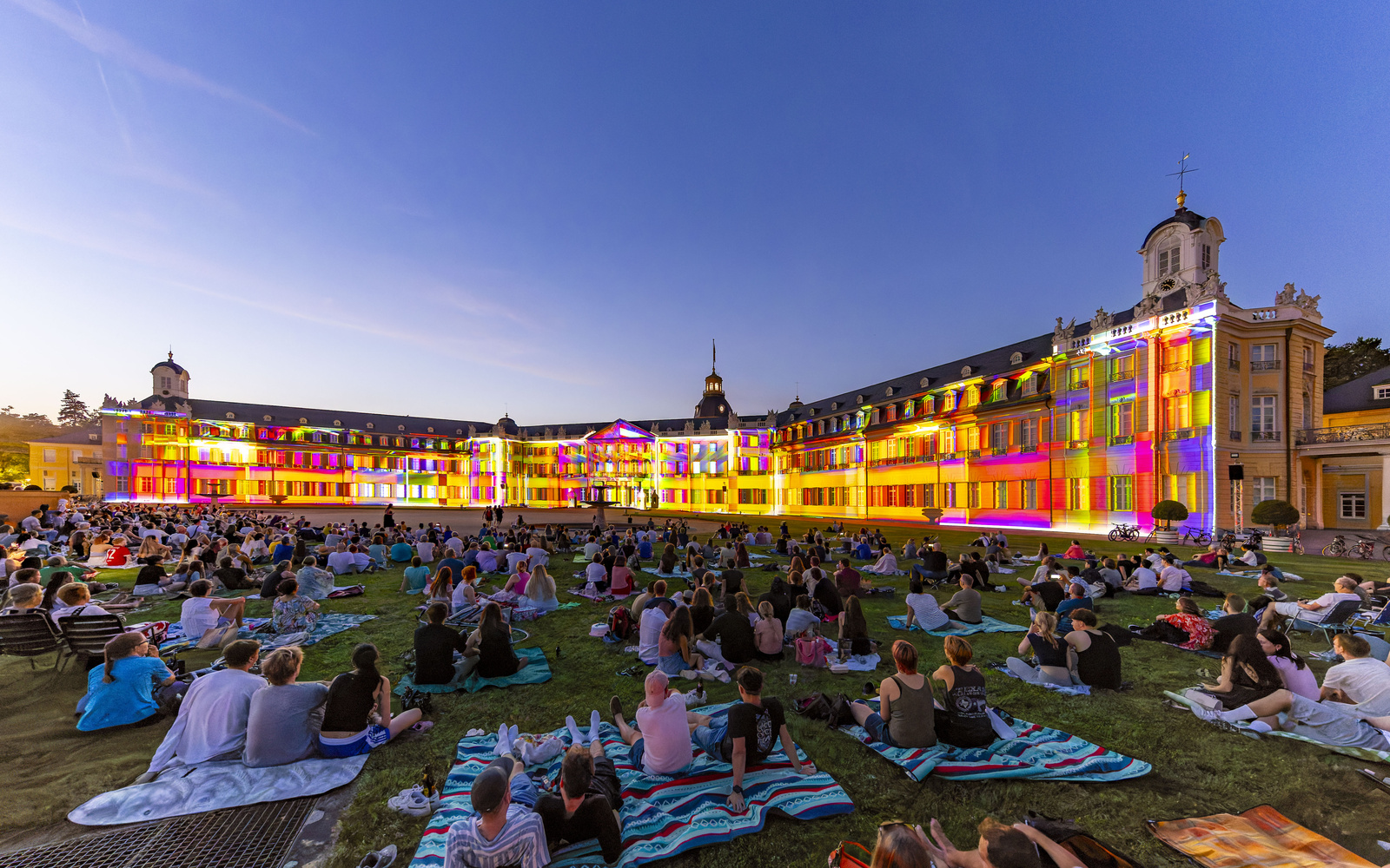 Personen sitzen abends auf Picknickdecken vor dem Karlsruher Schloss, das bunt angestrahlt wird.