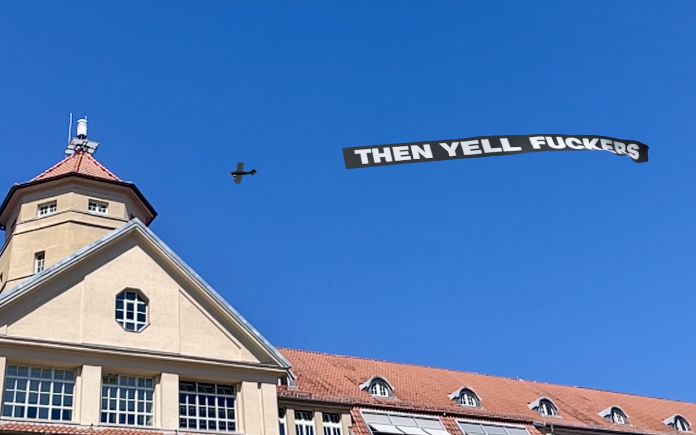 Zu sehen ist ein Flugzeug am Himmel über dem ZKM, das ein schwarzes Banner mit Schrift hinter sich herzieht.