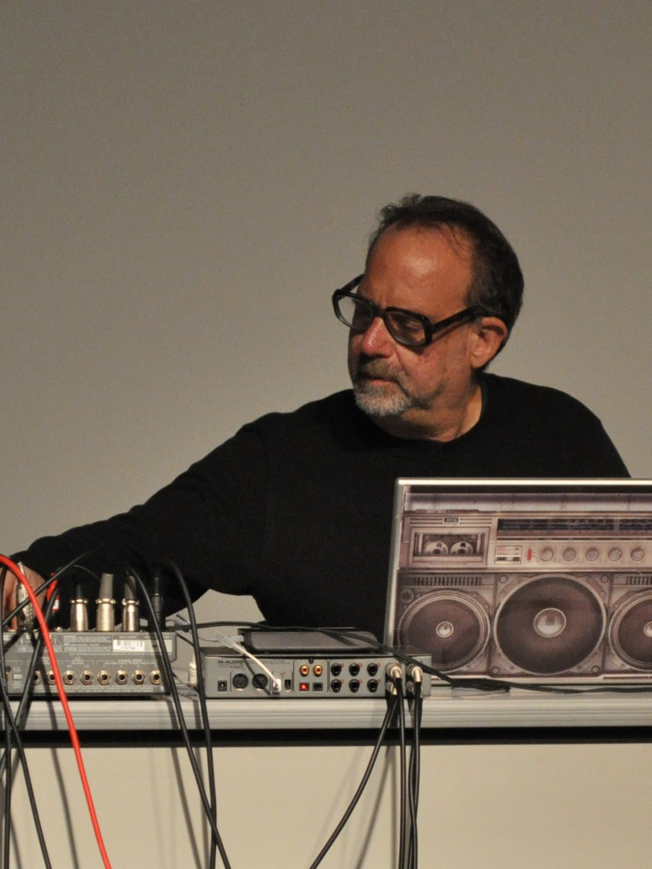 Man middle age, dressed in a black sweater and wearing black glasses, sits in front of a desk with a computer and some technical devices. He's looking down and creates live computer music.