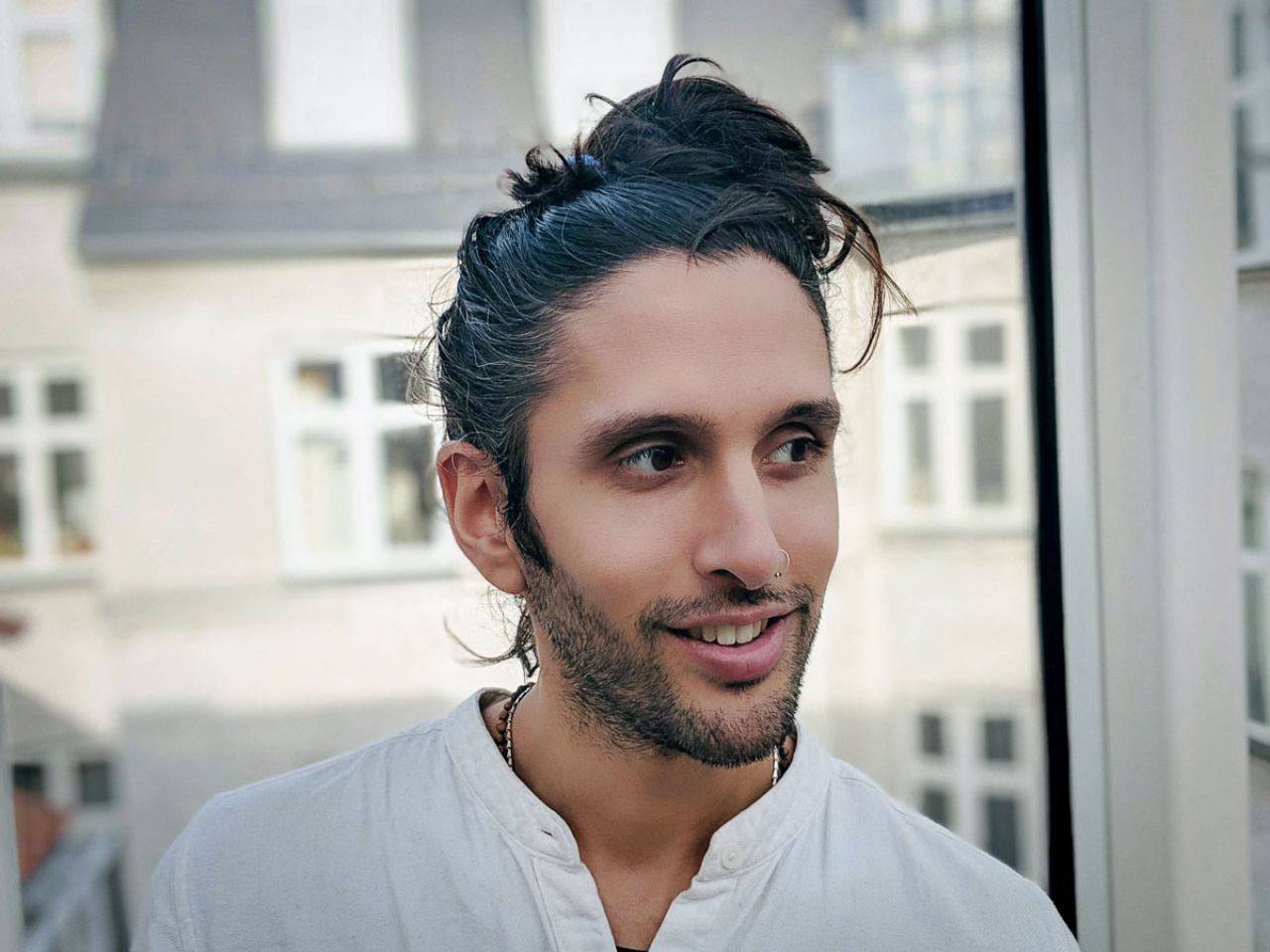 Auf dem Bild ist die Designerin und Künstler Cyrus Clarke zu sehen. Er trägt seine schwarzen langen Haare in einem Dutt und schaut leicht nach rechts unten.
