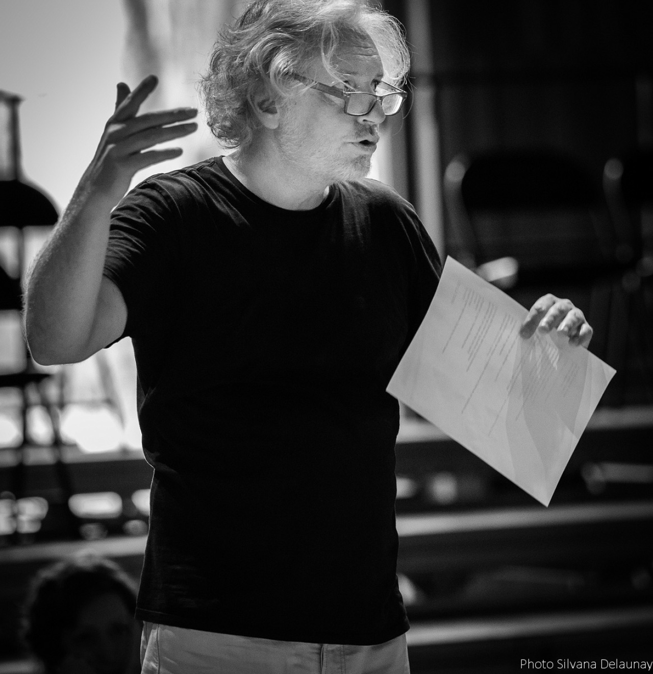 Black and white photo of Jean Pierre Seyvos, a man with curly hair and glasses, he is talking and holding a sheet of paper.