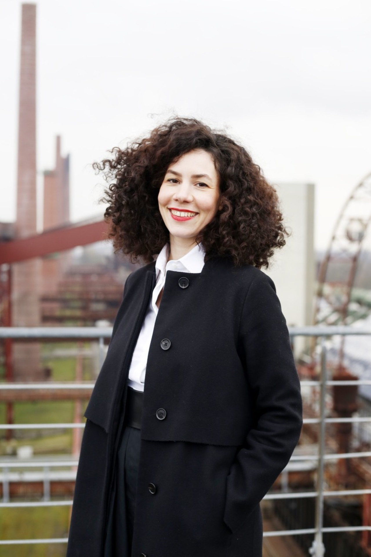 Jasmin Grimm in a black coat and white blouse standing outside in factory area