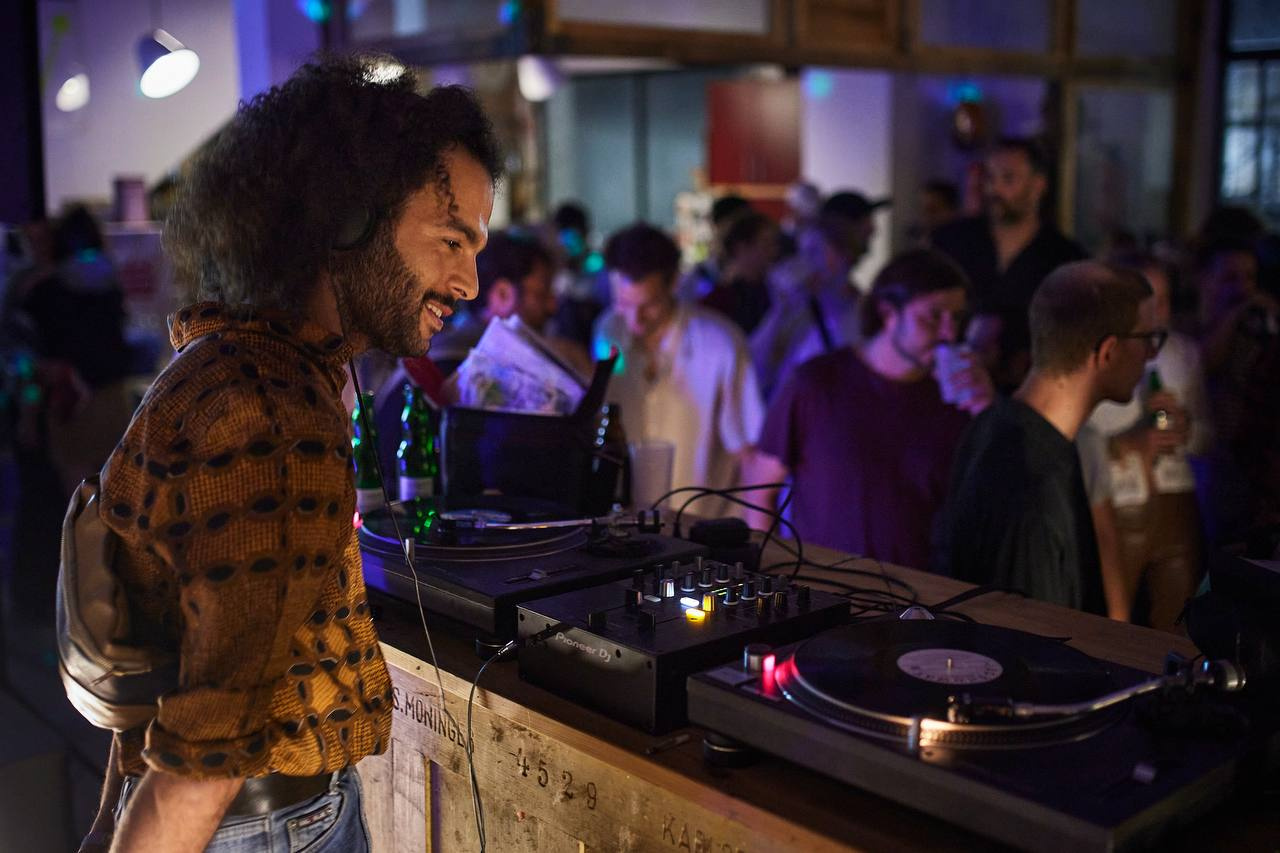 The picture shows a man in half-profile in front of a DJ booth, wearing a colorful shirt and black curls.