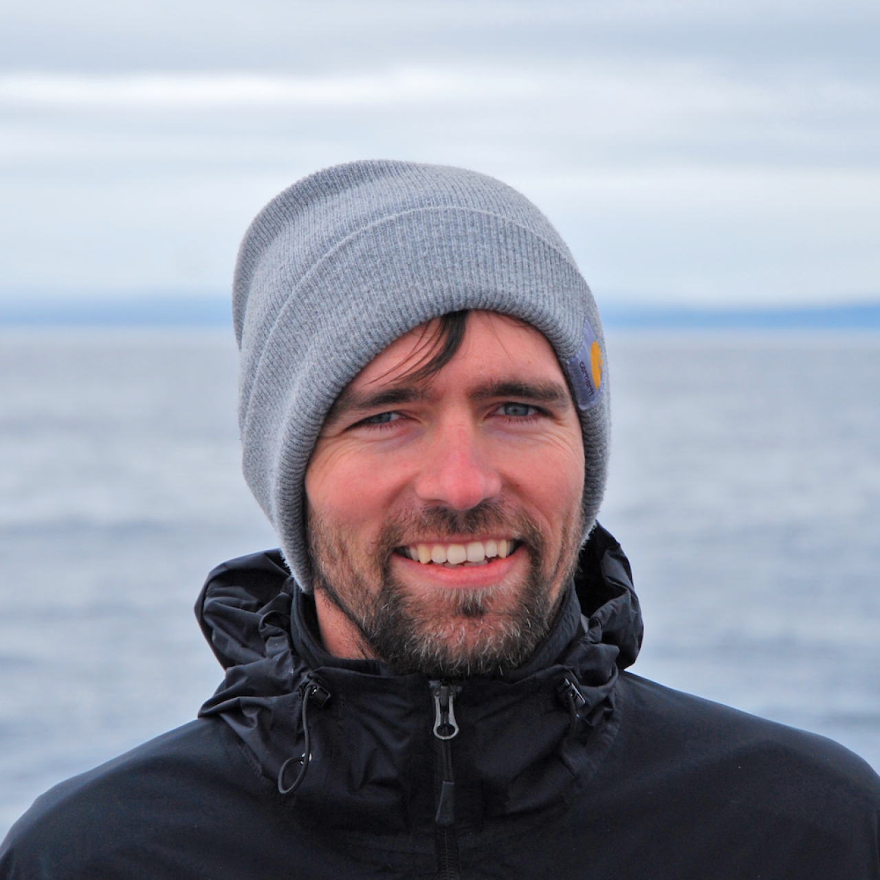 A man with a winter hat and rain jacket, the sea can be seen in the background.