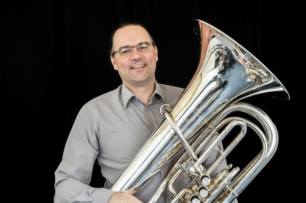 Zu sehen ist das Portraitfoto einer Person, die ein Musikinstrument hält. 