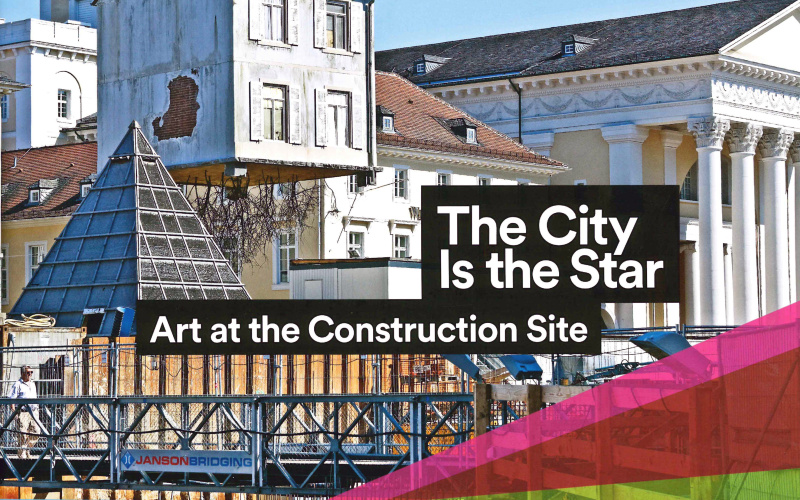 View of the Karlsruhe market place: A house is dangling from a crane. It has roots.