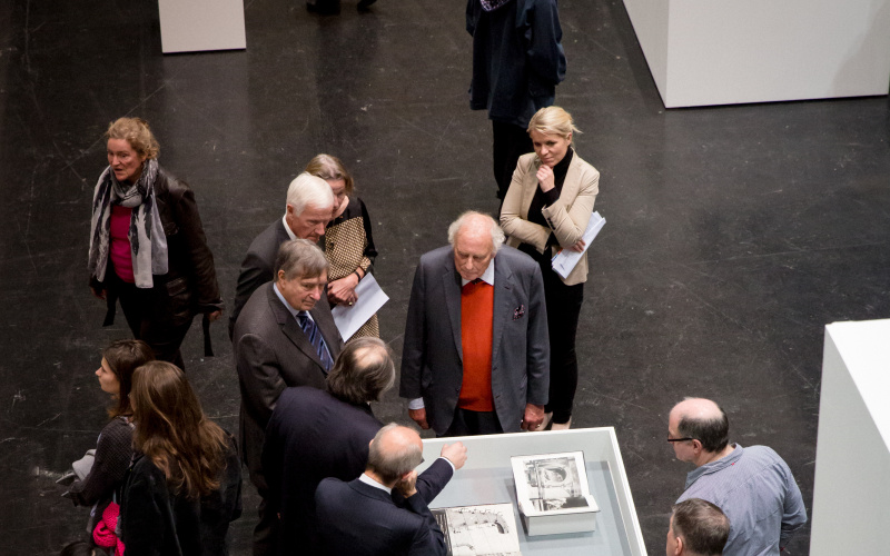 Die Besucher stehen um Herrn Weibel und eine weiße Vitrine, in der mehrere Bücher aufgeschlagen liegen.