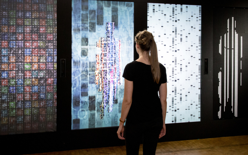 Eine Frau steht vor der Installation YOU:R:CODE, einer Mehrkanalprojektion, die sie in Datenform darstellt