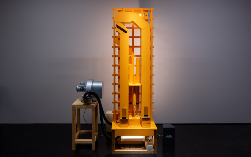 Zu sehen ist eine rechteckige, maschinenartige Installation, welche auf einem Hocker steht. Links neben der Installation steht ebenfalls ein kleiner Hocker, auf welchem ein Objekt liegt. 