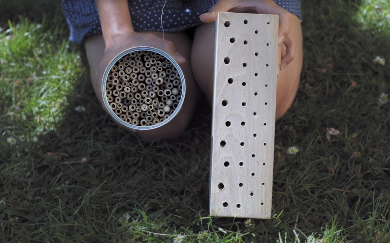 Zwei verschieden gefertigte Nisthilfen, eine aus einem Klotz holz und die anderen aus einer Dose gefüllt mit Strohhalmen, die sich besonders für Wildbienen eignen werden in die Kamera gehalten.