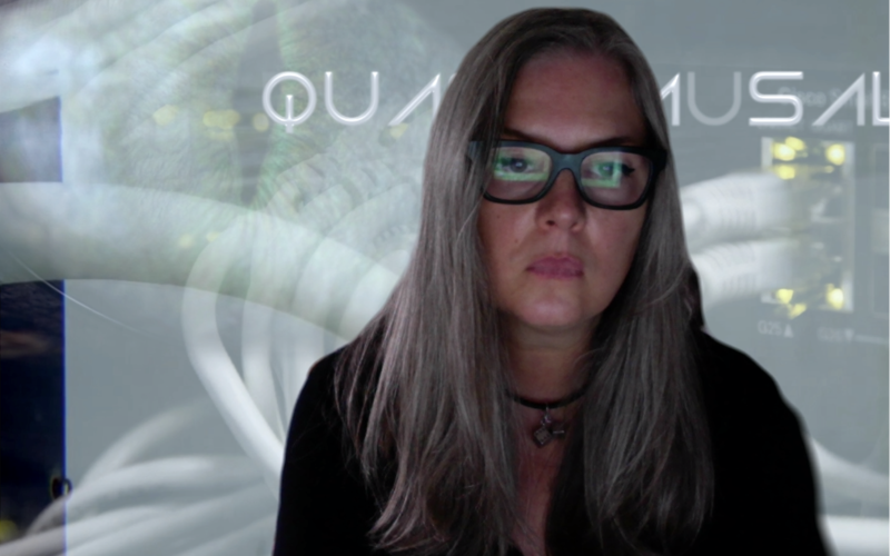 Ami Clarke, a woman with long hair and glasses with a thick, square frame, looks at a screen. This screen is reflected in her glasses.