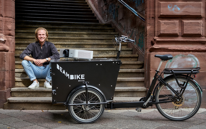 Das Foto zeigt einen Mann, der auf einer Treppe sitzt. Vor ihm steht ein Fahrrad an dem ein Beamer befestigt ist, das »beambike«.