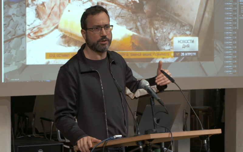 Eyal Weizman, ein Mann mit Bart und Brille hält einen Vortrag vor einer Leinwand..