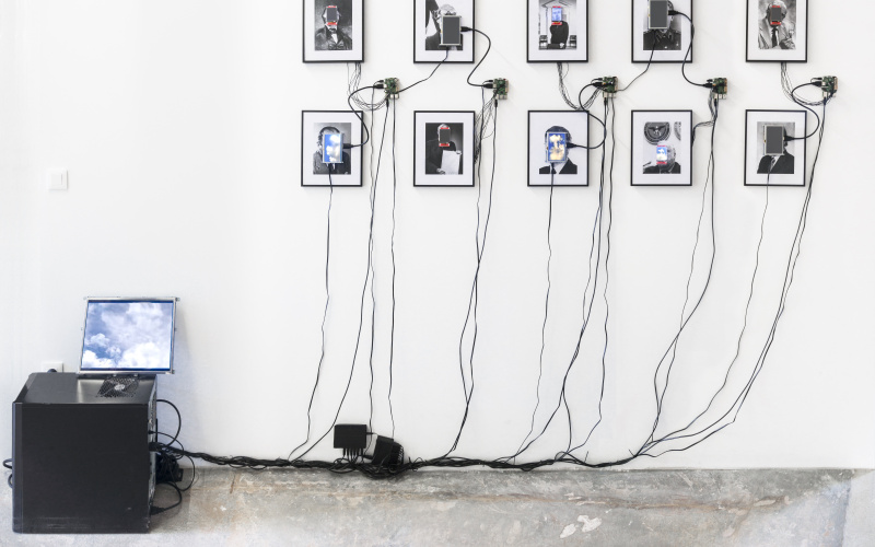 Das Foto zeigt 10 eingerahmte Portraits auf deren Gesichter kleine Monitore angebracht sind. Diese sind durch Kabel mit einem Computer verbunden.