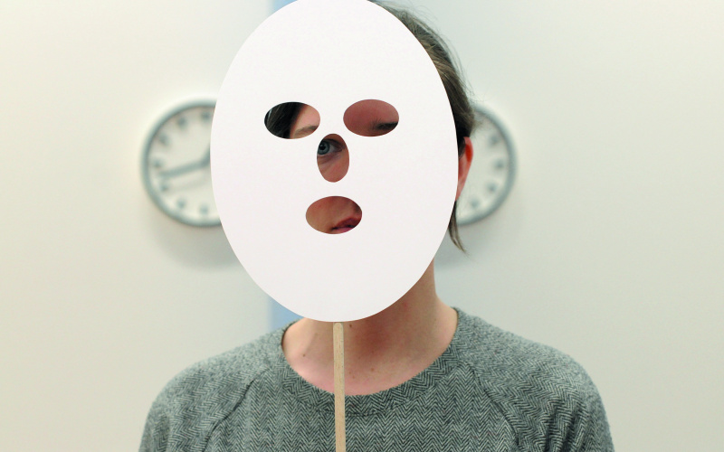 Eine Person undefinierten Geschlechts hält sich eine weiße Papiermaske vors Gesicht.