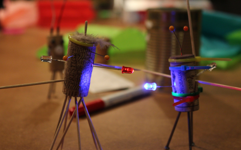 Zwei Korken, die zu kleinen Figuren umgewandelt wurden und mit LEDs ausgestattet sind stehen auf einem Tisch mit allerlei Bastelutensilien.