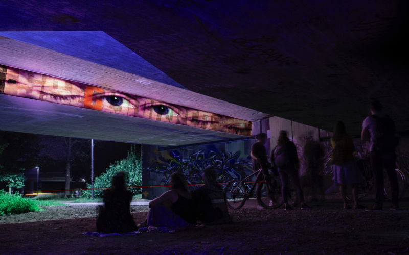 Eine Brücke in einem Park bei Nacht. An die Außenwand der Brücke ist in einem horizontalen Streifen eine Reihe Augenpaare projiziert. Der Rets der Brücke leuchtet in verschiedenen Farben.
