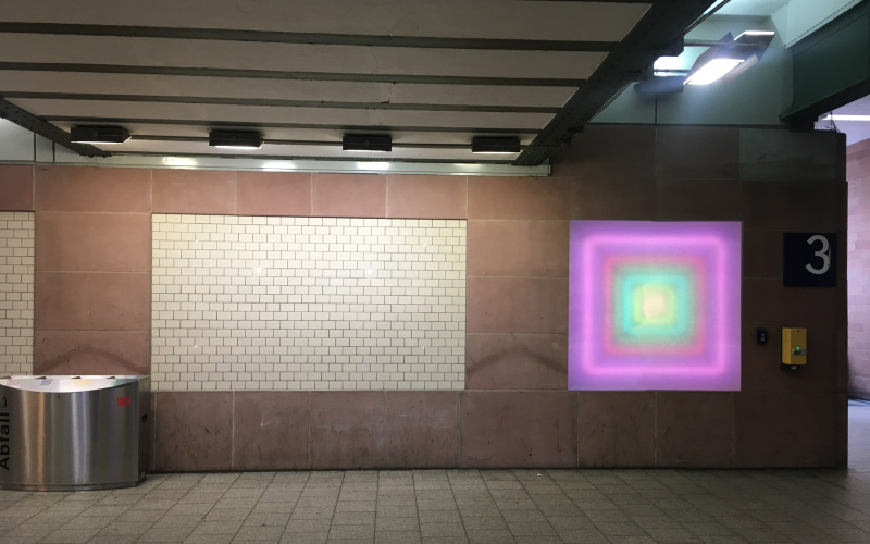 Zu sehen ist der Durchgang unter dem Karlsruher Hauptbahnhof. An der Wand ist ein leuchtendes Quadrat angebracht, das von innen nach außen einen Farbverlauf hat, der eine Tiefe erzeugt.