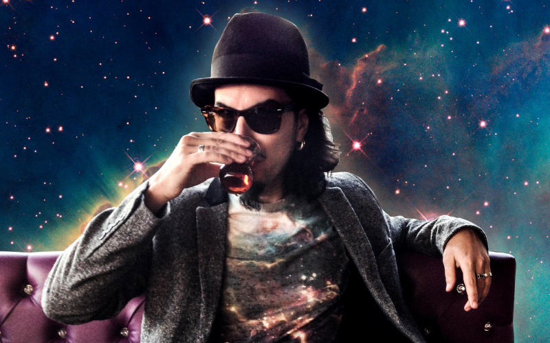 Memo Aktens is sitting on a violet velvet couch and in front of a backgammon game, drinking turkish tea. The background is a colage of the universe. 