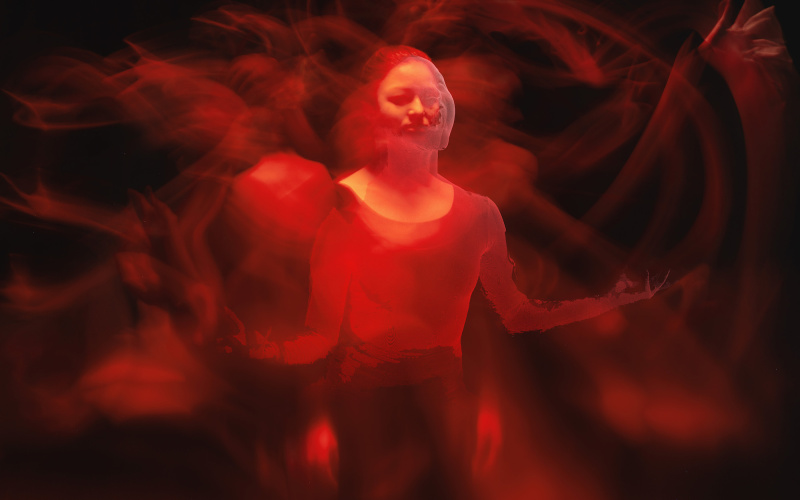 Das Foto zeigt eine von rotem Licht erleuchtete Frau in einem abgedunkelten Raum. In dieser Impression sieht man mehrere verschwommene Arme und Hände in Bewegung, die fast einer indischen Gottheit ähneln. 