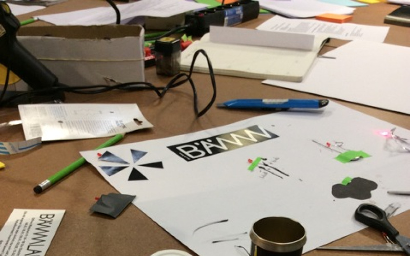 Multiple items for crafting are lying on a table such as batteries, electronics, paper or tools. Amongs that a piece of paper, on which some LEDs are taped on.