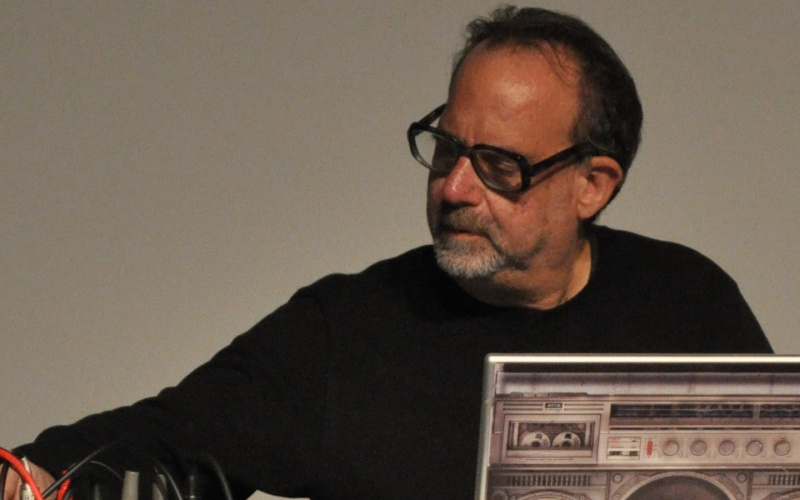 Man middle age, dressed in a black sweater and wearing black glasses, sits in front of a desk with a computer and some technical devices. He's looking down and creates live computer music.