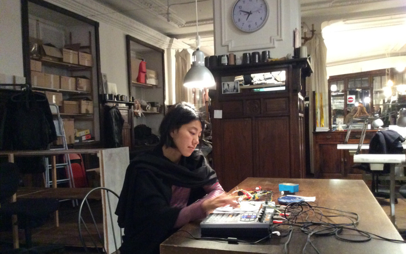 A young woman is sitting at a table in front of some technical devices. She's creating electronic music.