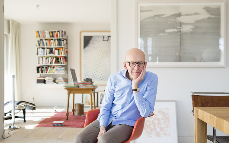 Professor Bude sits on a red chair in an apartment with bookshelf and desk, his head resting on his hand.