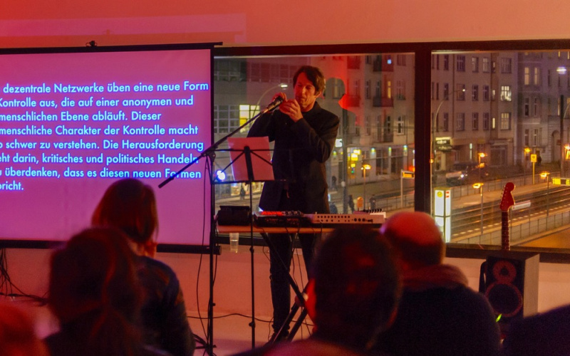 Ein Mann bei seiner Performance am Keyboard und hinter ihm eine Leinwand, auf die ein Text projiziert ist.
