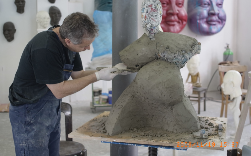 Ein Mann in Latzhosen modelliert eine voluminöse Figur aus Ton.