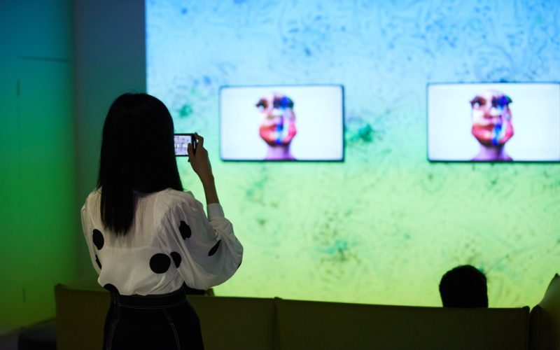 Das Foto zeigt die Arbeit »im here to learn so :))))))«. Im Hintergrund ist eine große Leinwand auf der ein Google DeepDream projiziert ist. Drei davor liegende Bildschirme zeigen den Kopf des Chatbots Tay.