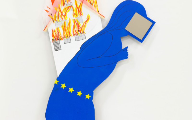 Eine gebückte in blau vermummte Figur mit einem Screen vor dem Gesicht trägt auf ihren Schultern ein brennendes Haus. Am Rand der blauen Kutte und auf Hüfthöhe sind Sterne eingewebt.