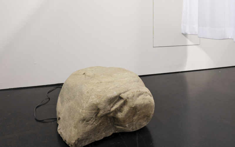 Zu sehen ist ein großer hellgrauu/beiger Stein, der mit einem Kabel verbunden ist. Hinter ihm hängen an der Wand zwei gerahmte Bilder und ein Krankenhausvorhang ist leicht angeschnitten zu sehen.