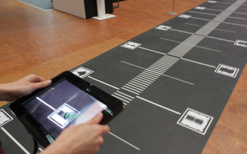 Ausschnitt eines Weges voll mit QR-Codes. Eine Hand, die mit einem iPad die QR-Codes abscannt