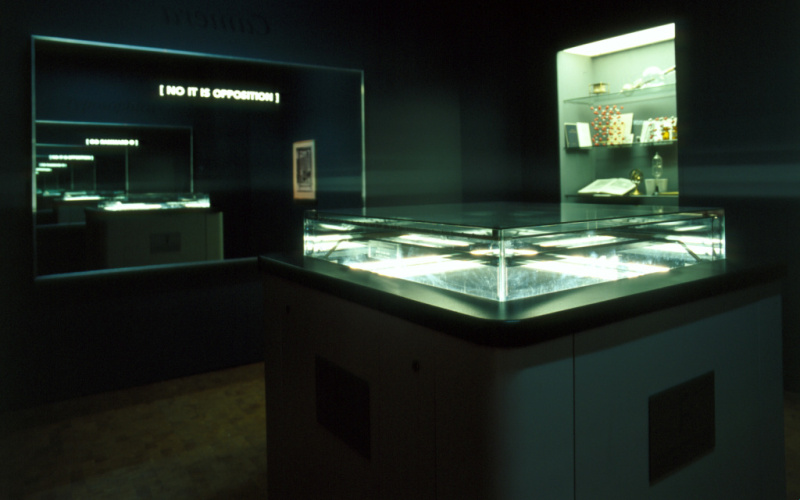 Der Blick in die Ausstellung »Iconoclash« zeigt "the typosophic pavilion"  von Ecke Bonk  und Richard Hamilton: einen dunklen Raum mit erleuchteten Vitrinen und einem Spiegel mit der Aufschrift »No It Is Opposition«