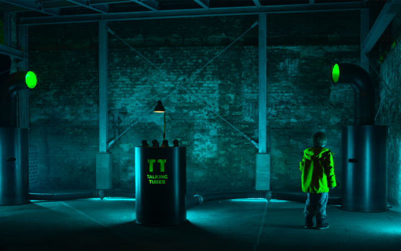 Ein blau erleuchteter Raum mit einem Kind, das die »Talking-Tubes«-Installation betrachtet: ein Steuerungspult zwischen zwei großen Röhren